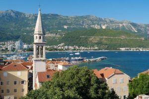 Church spire rises above beige buildings in Budka Montenegro with harbor, boats and high green cliffs in background