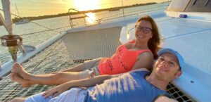 A gal and guy who took the Offshore Sailing School cruising course are relaxing on front of a big catamaran with glow of the sun in the distance