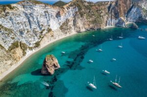 Aerial shot of boats anchored in clear green water with large rock formations and beach off Ponza Island in Sicily