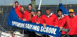 The Barr family of 4 gals and 4 guys all in matching red jackets leaning on the boom of an Offshore Sailing School Colgate 26