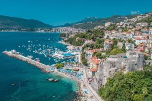 Aerial shot of Herceg Novi in Montenegro showing lots of boats anchored, big swimming pool and new and ancient buildings on right shore